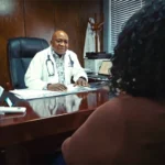 Dr. Songabau, wearing his white coat and stethoscope, seated at his desk having a consultation with a patient in his office.